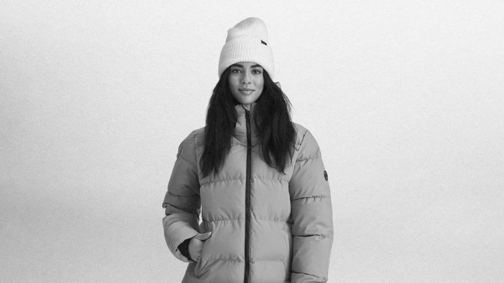 A person wearing a puffer jacket and knit hat, standing facing forward with hands in pockets, against a plain background.